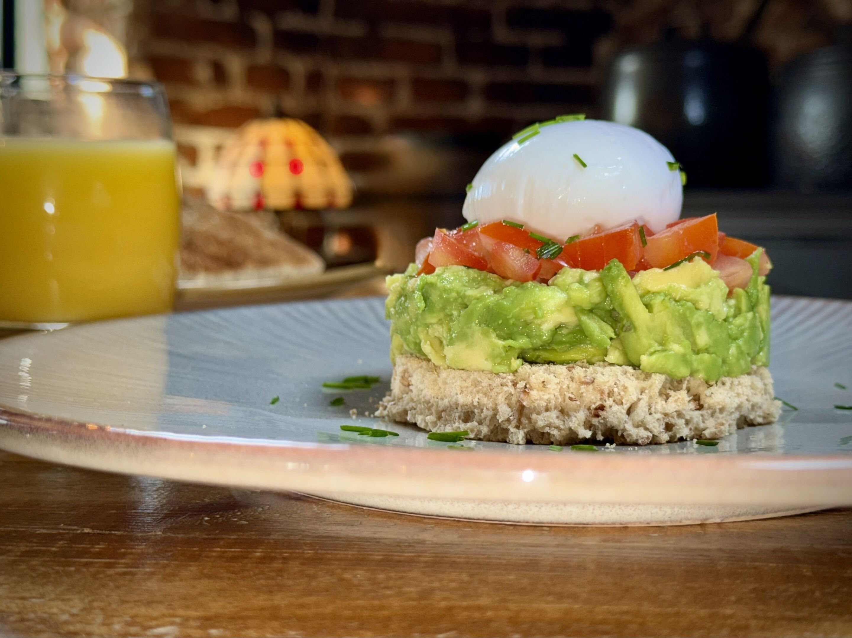 Un plato con un sándwich cubierto de aguacate, tomate y un huevo pochado, con un vaso de jugo de naranja al fondo.