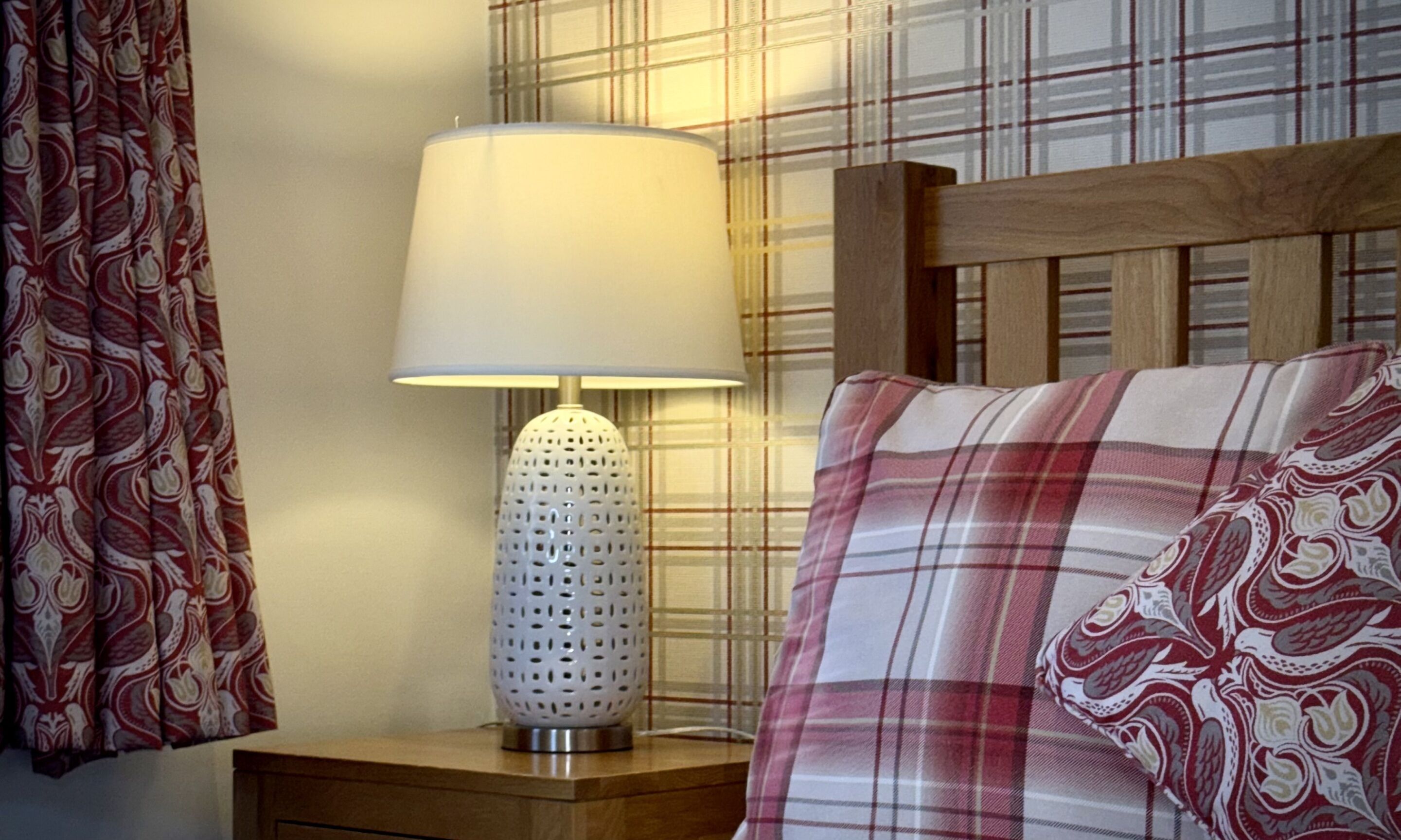 A bed with a plaid pillow and a white lamp on a wooden nightstand.