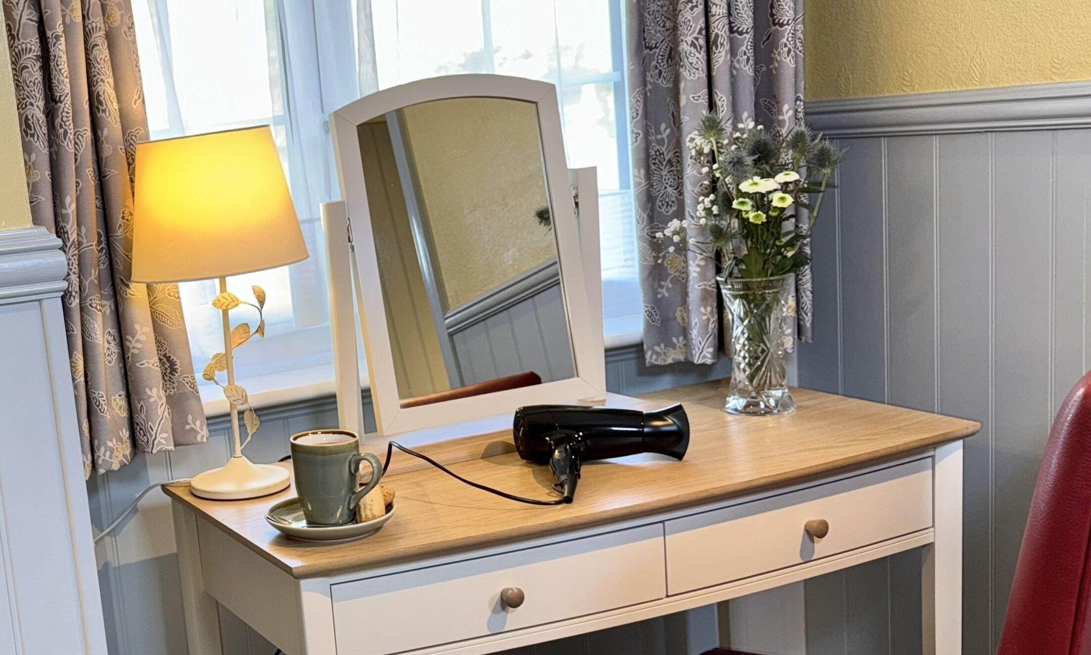 A vanity with a mirror, a lamp, a cup, and a vase with flowers.