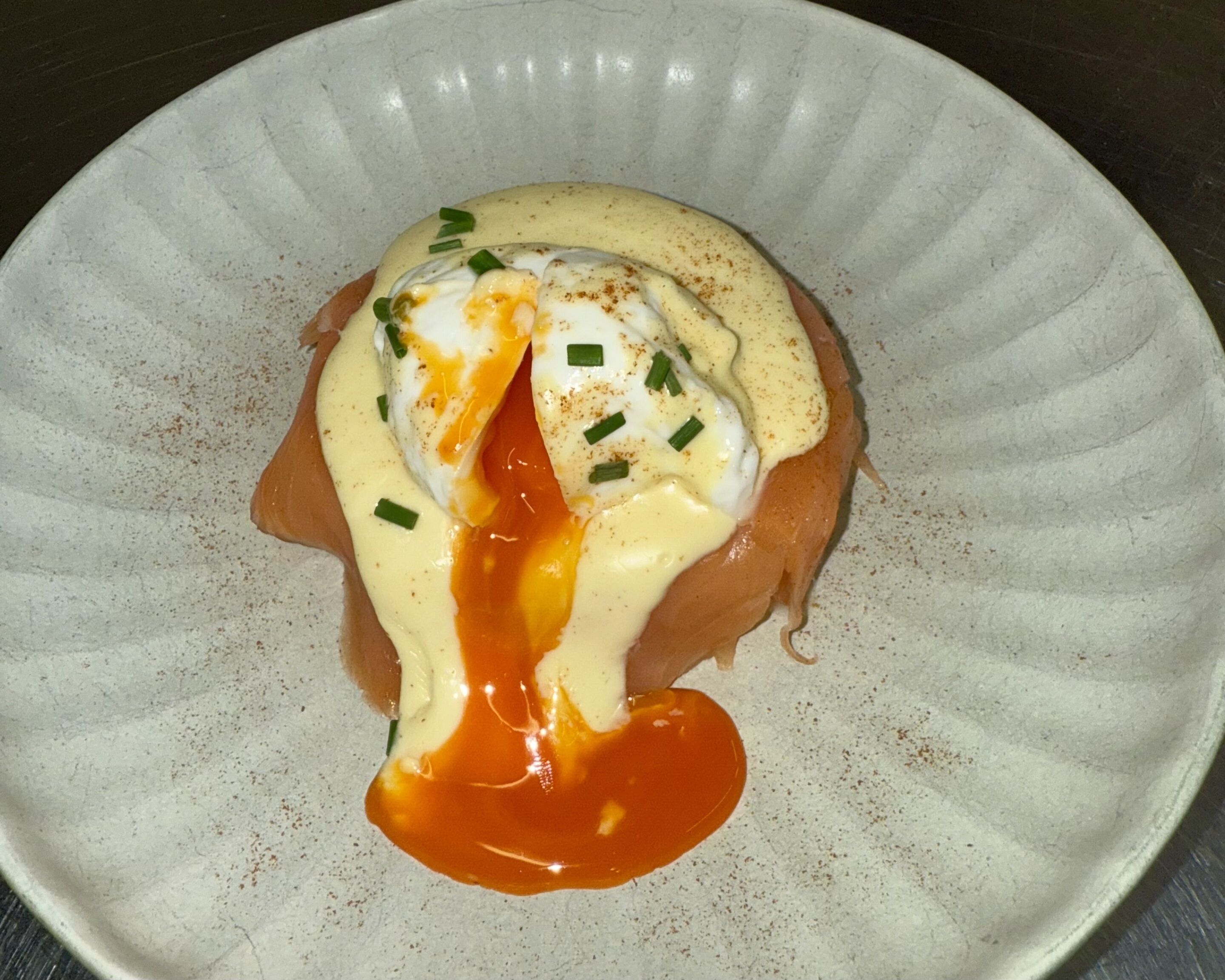 Une assiette en céramique blanche sur laquelle on a disposé un œuf poché, une sauce hollandaise et de la ciboulette.