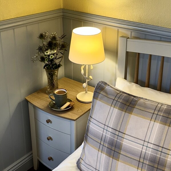 A neatly made bed with a plaid pillow, a bedside table with a lamp, a cup of coffee, and a vase with flowers. - Sibton White Horse Inn (Saxmundham)