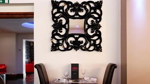 A hotel room with a dining table, two chairs, a decorative mirror, and a small table with a wine glass and a menu. - Ashiana Hotel (Newark)