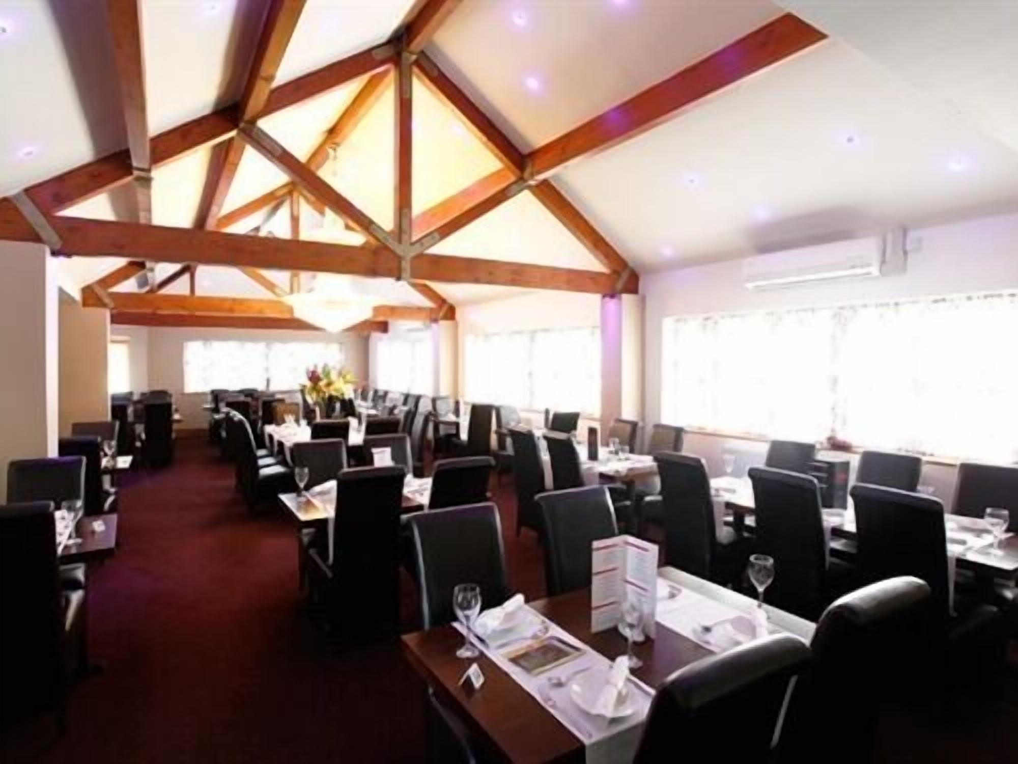 A dining area with high wooden beams, tables set with chairs, and large windows allowing natural light.