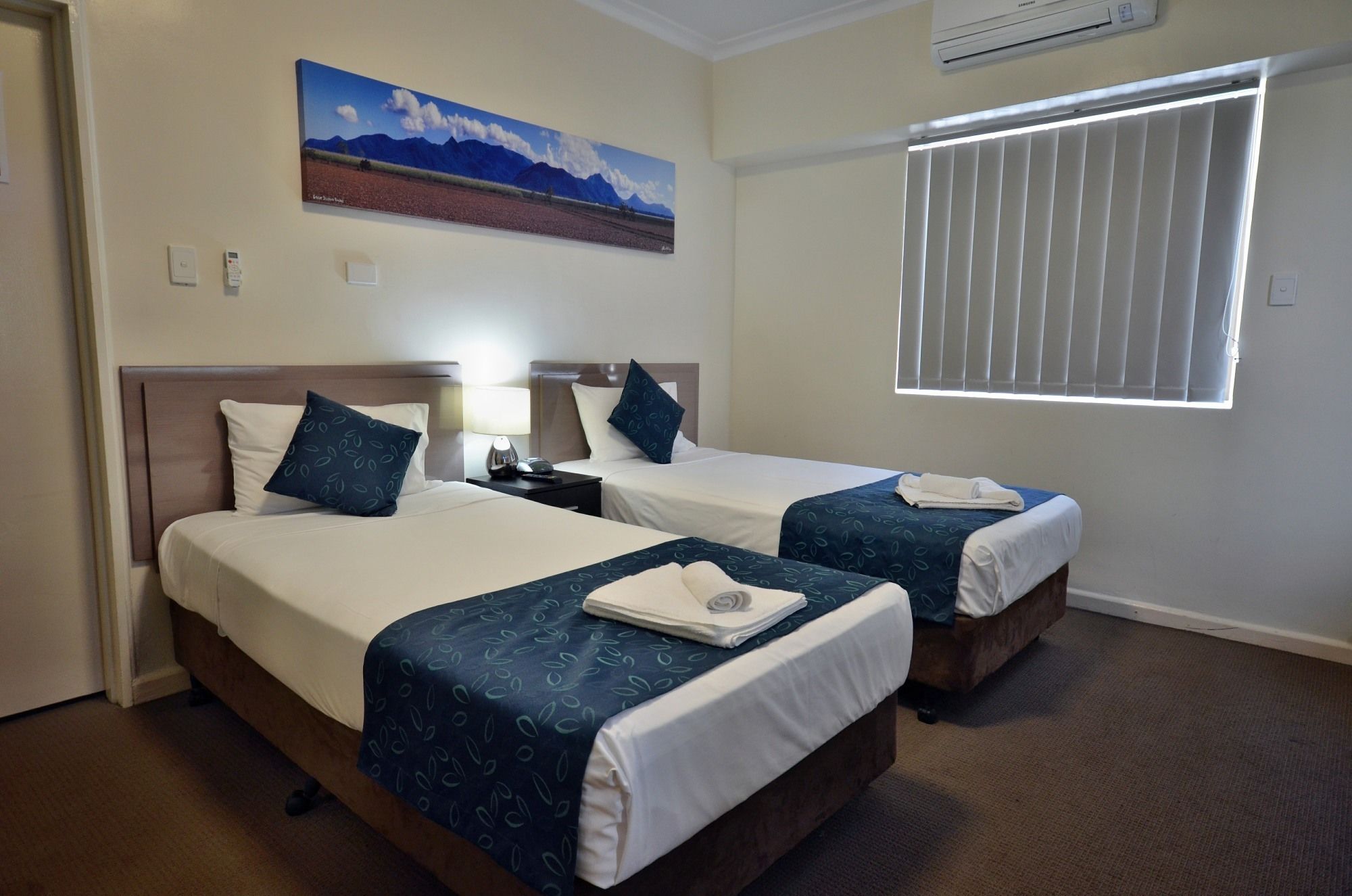 Quarto de hotel com duas camas, um quadro de montanhas, ar condicionado e uma janela com persianas.