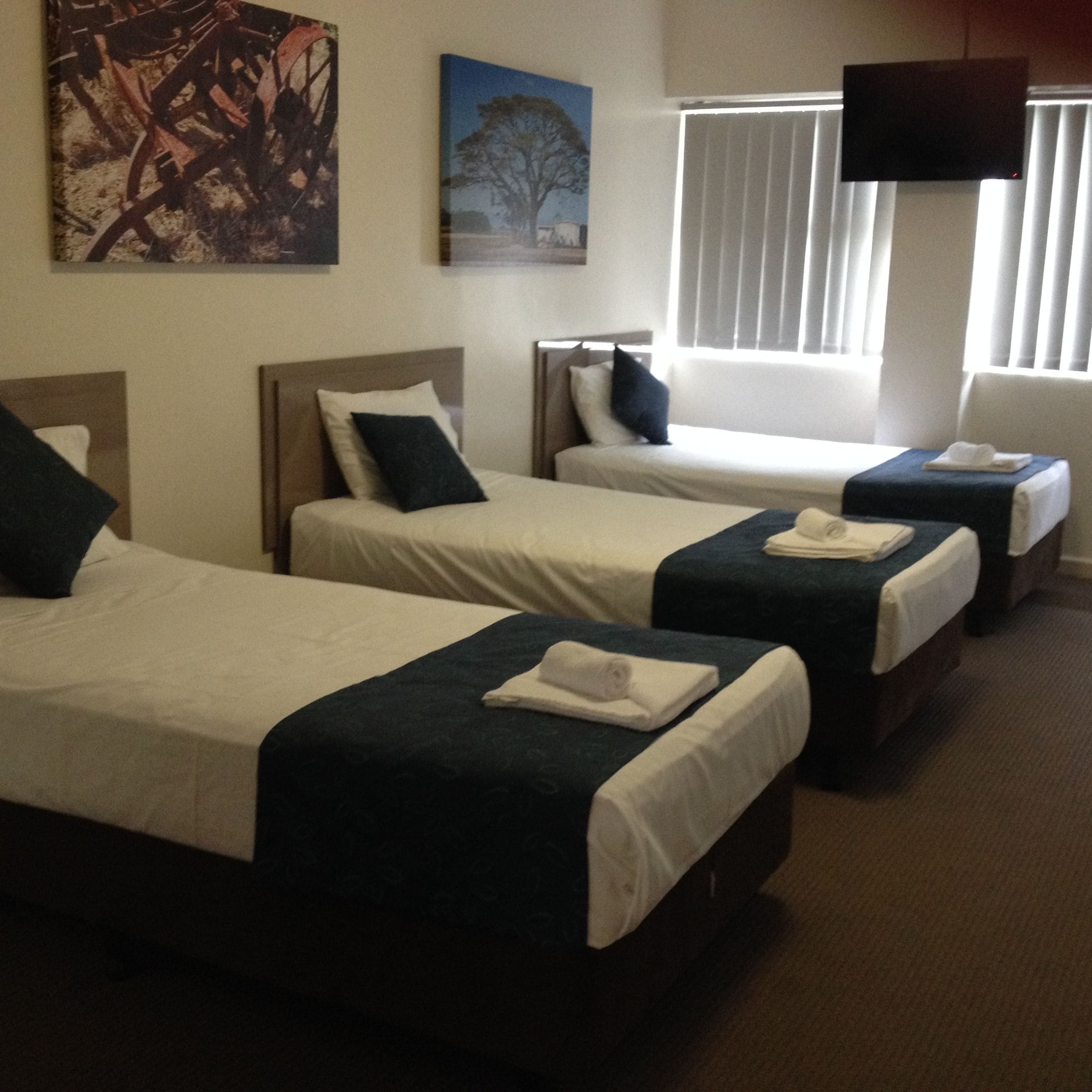 A hotel room with three single beds, each with a pillow and a rolled towel. There is a television mounted on the wall and a window with blinds.
