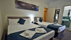 Habitación de hotel con dos camas, cada una con cabecera, un cuadro sobre las camas, una mesita de noche con lámpara, y un baño visible a través de una puerta abierta.
