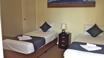 A hotel room with two beds, each with a pillow and a folded towel. There is a nightstand with a lamp and a phone between the beds. A painting of a landscape is on the wall above the beds.