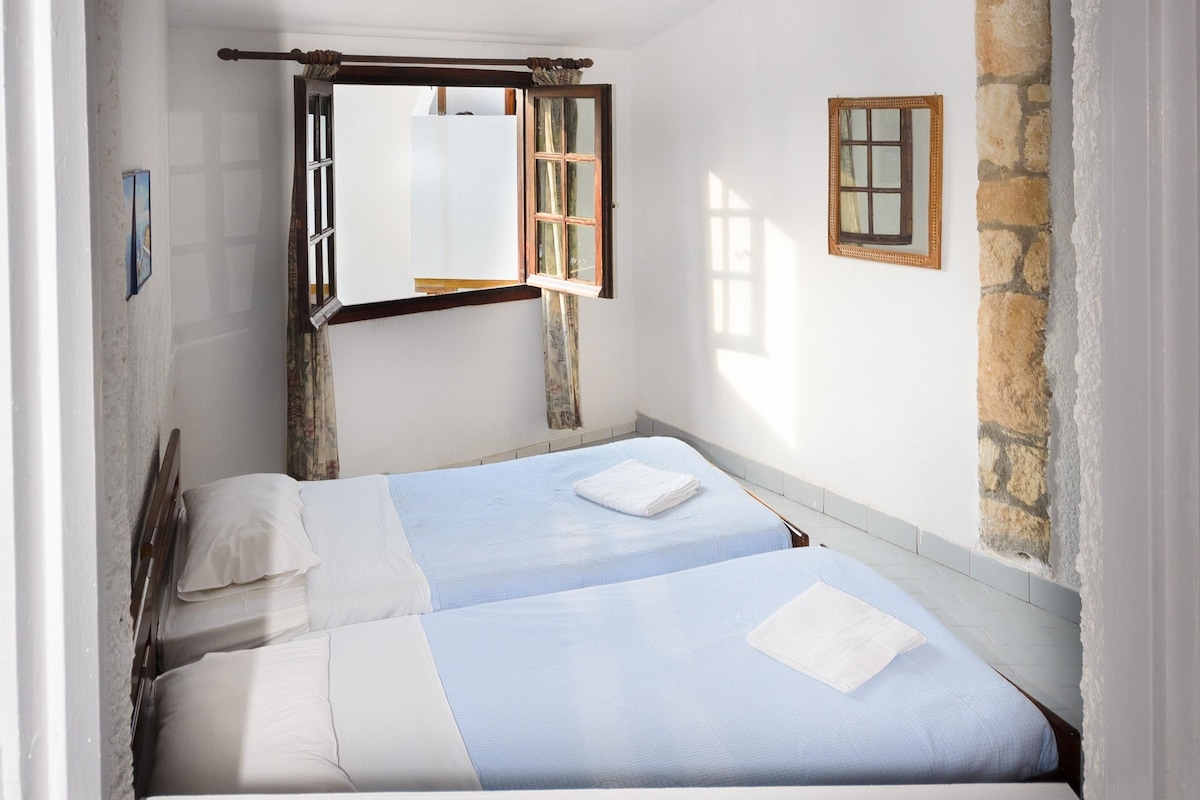 A small, well-lit bedroom with two beds, a window with a wooden frame, a mirror, and a stone wall.