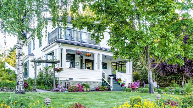 Una casa de dos pisos con un porche, rodeada de un jardín bien cuidado con diversas flores y arbustos.