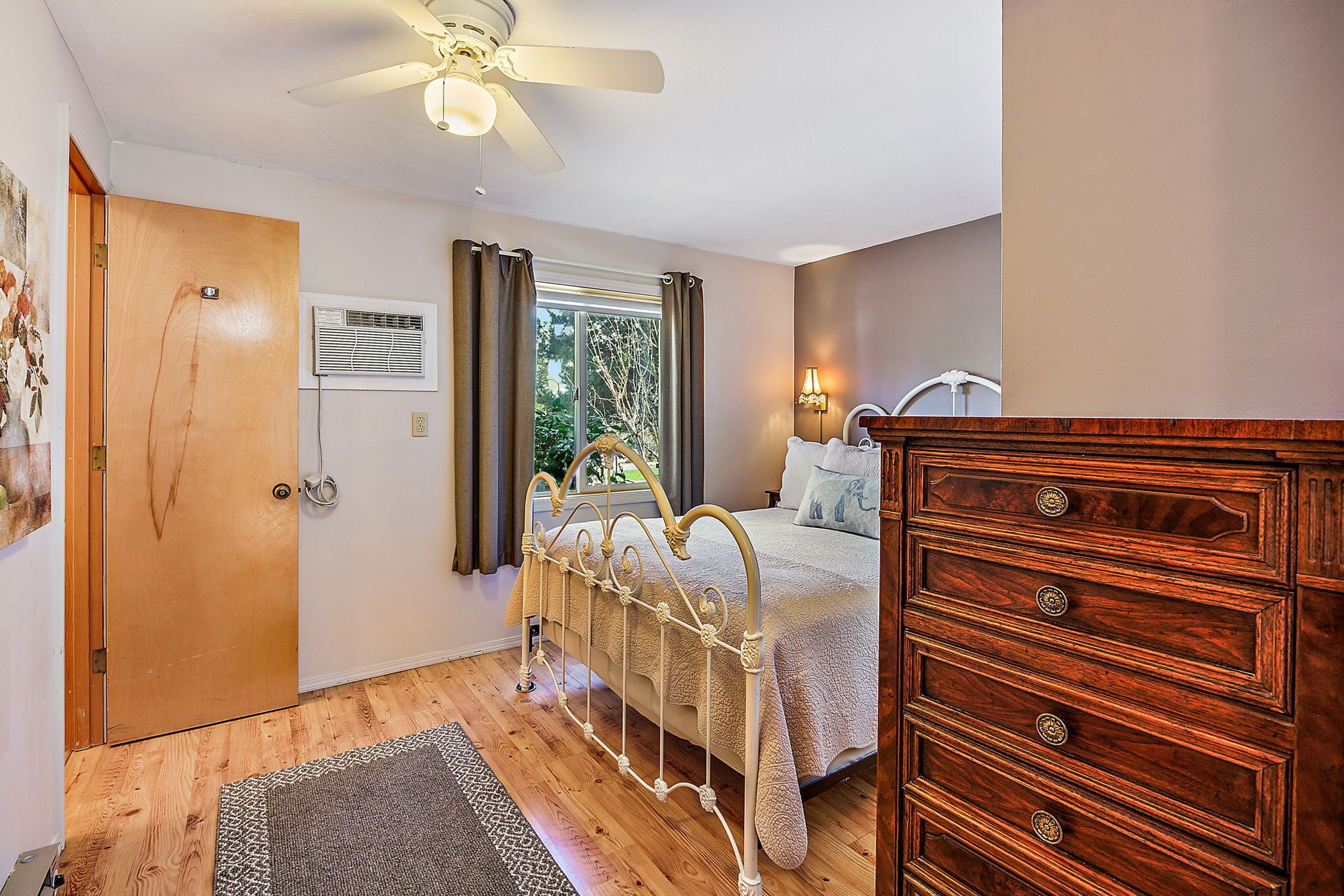 A bedroom with a bed, wooden dresser, ceiling fan, window with curtains, and a wall-mounted air conditioning unit.