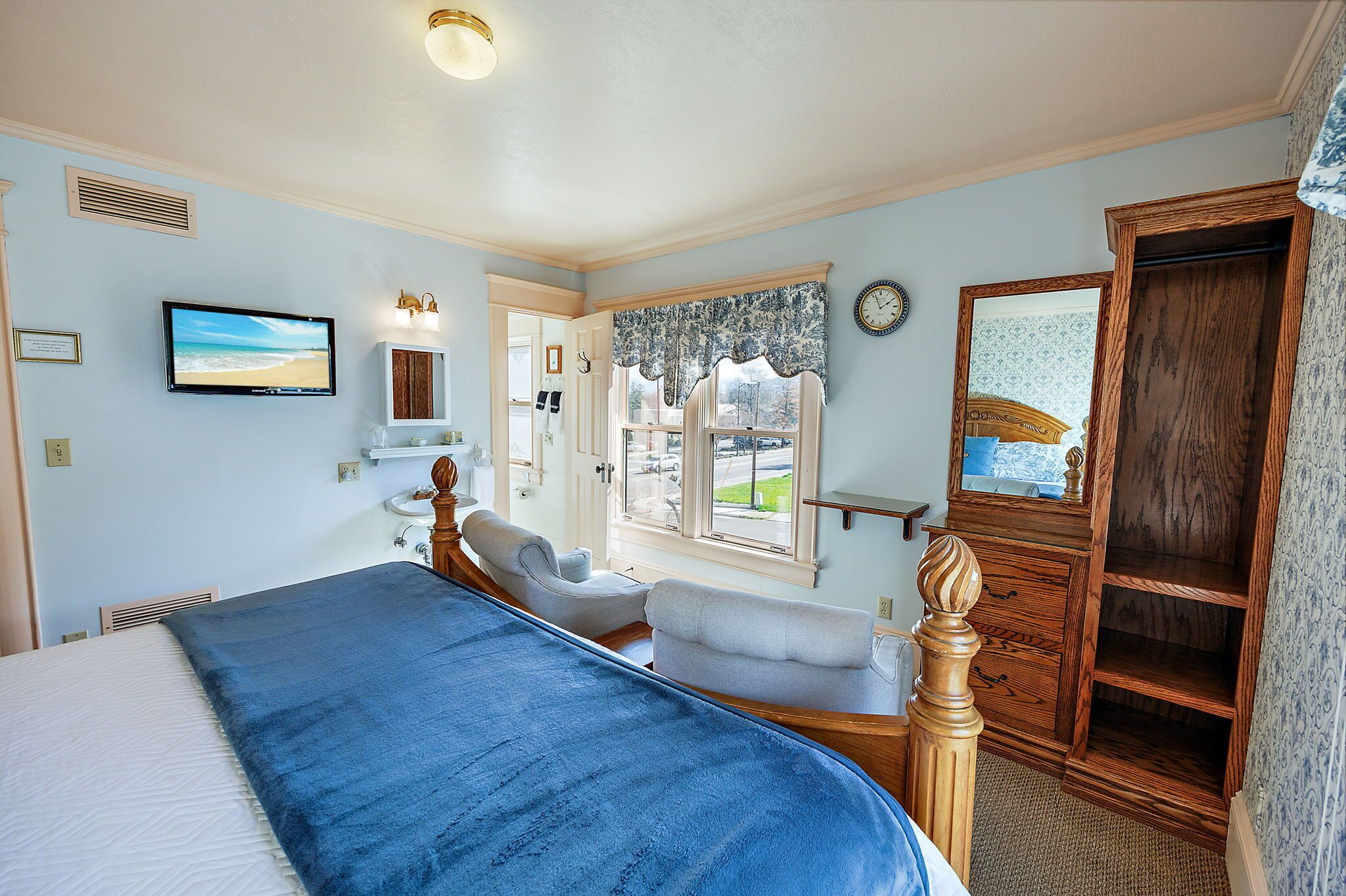 A bedroom with a bed, two chairs, a dresser, a mirror, a clock, and a window with a view of the outdoors.