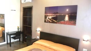 A hotel room with a bed, a desk, and a chair. There is a picture on the wall and an air conditioning unit above the desk. - B&B Il Campanile (Naples)