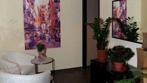 A modern living room with a white armchair, a small round table with a plant, and two potted plants. There is a large abstract painting on the wall and a mirror reflecting the painting. - B&B Il Campanile (Naples)