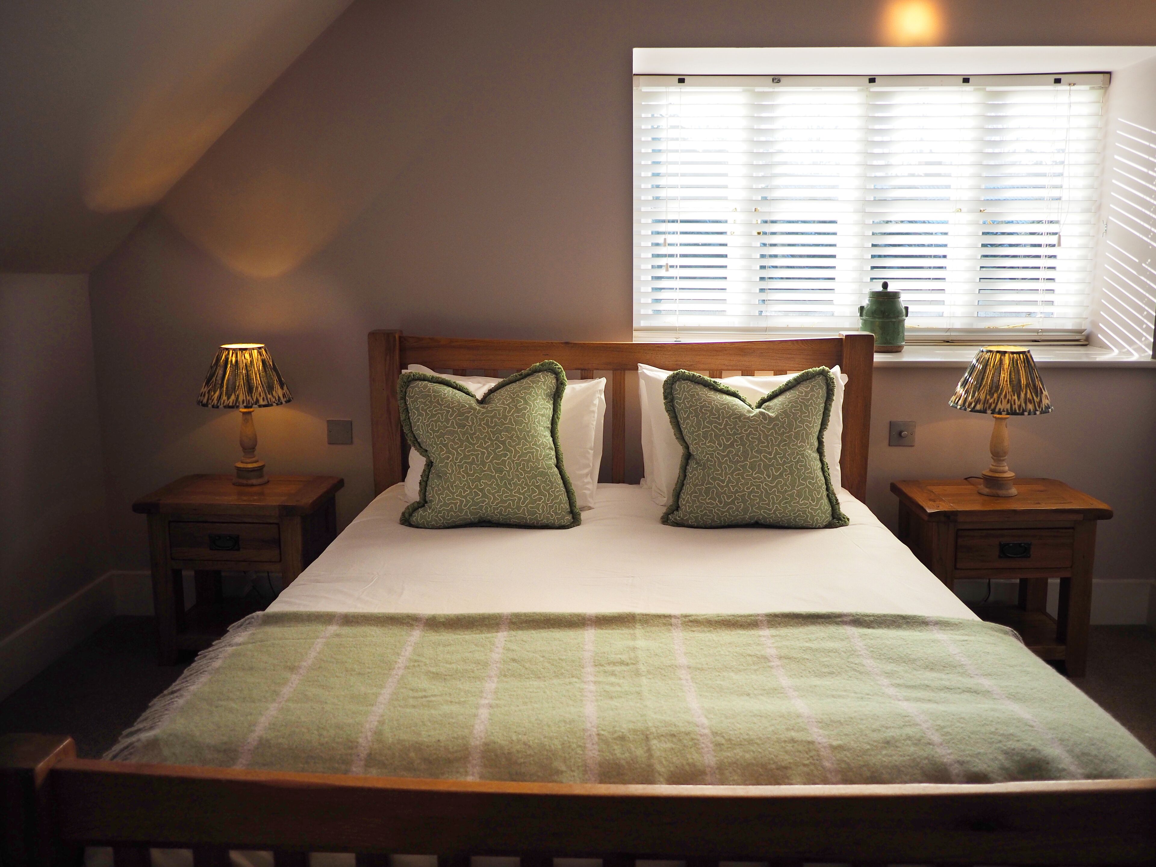 A bedroom with a bed, two bedside tables, two lamps, a window with blinds, and a green throw blanket.