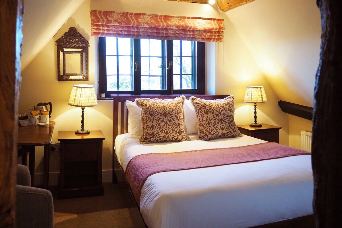 A cozy bedroom with a large bed, two bedside tables with lamps, a window with curtains, and a stone wall.