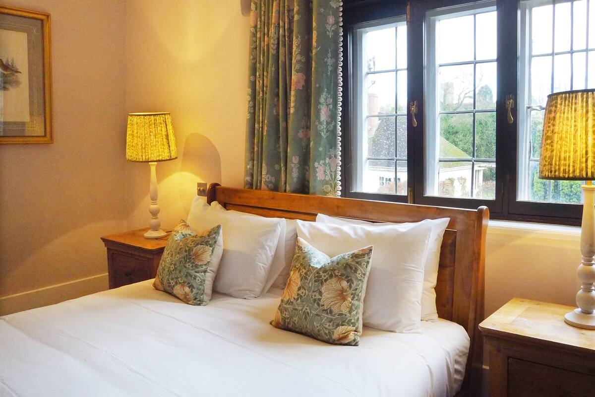 A bedroom with a wooden bed, floral pillows, a nightstand with a lamp, and a window with curtains.