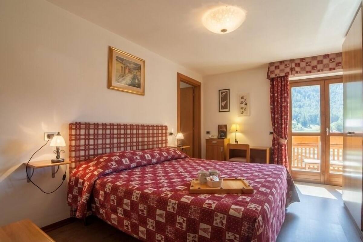 Un dormitorio con una cama, una mesita de noche, una lámpara, un ventanal con cortinas y un cuadro en la pared.