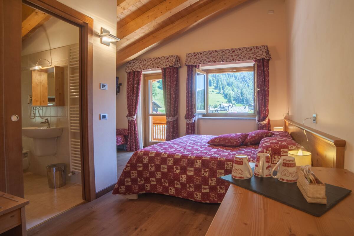 Un dormitorio con techo de madera, dos ventanas con cortinas florales, una cama con colcha estampada y una mesita de noche con tazas y una lámpara.