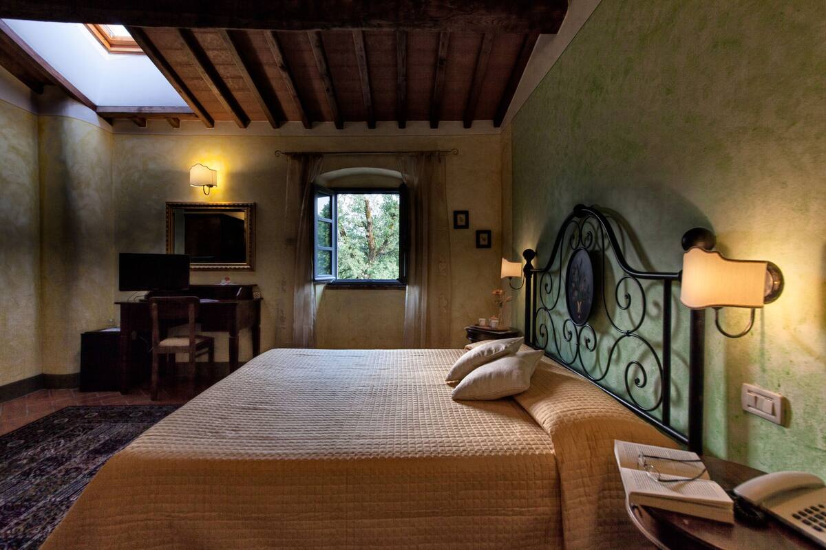 A bedroom with a bed, a desk, a chair, a TV, and a window with a view of trees.