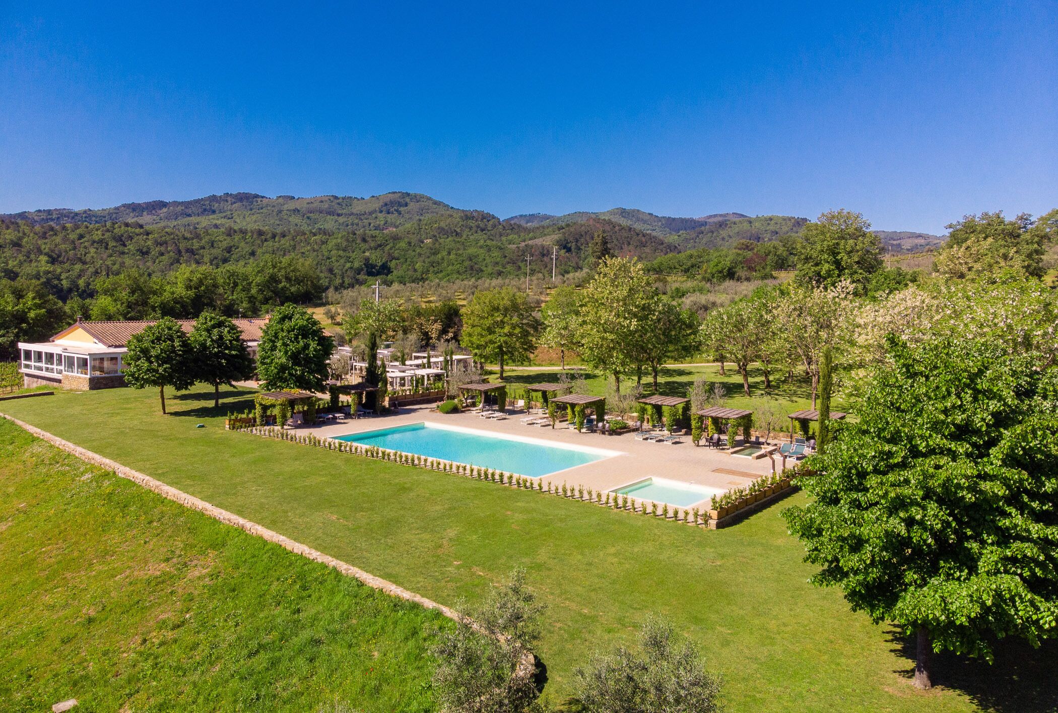 A large rectangular pool surrounded by lounge areas and covered seating, set against a backdrop of green hills and clear skies.