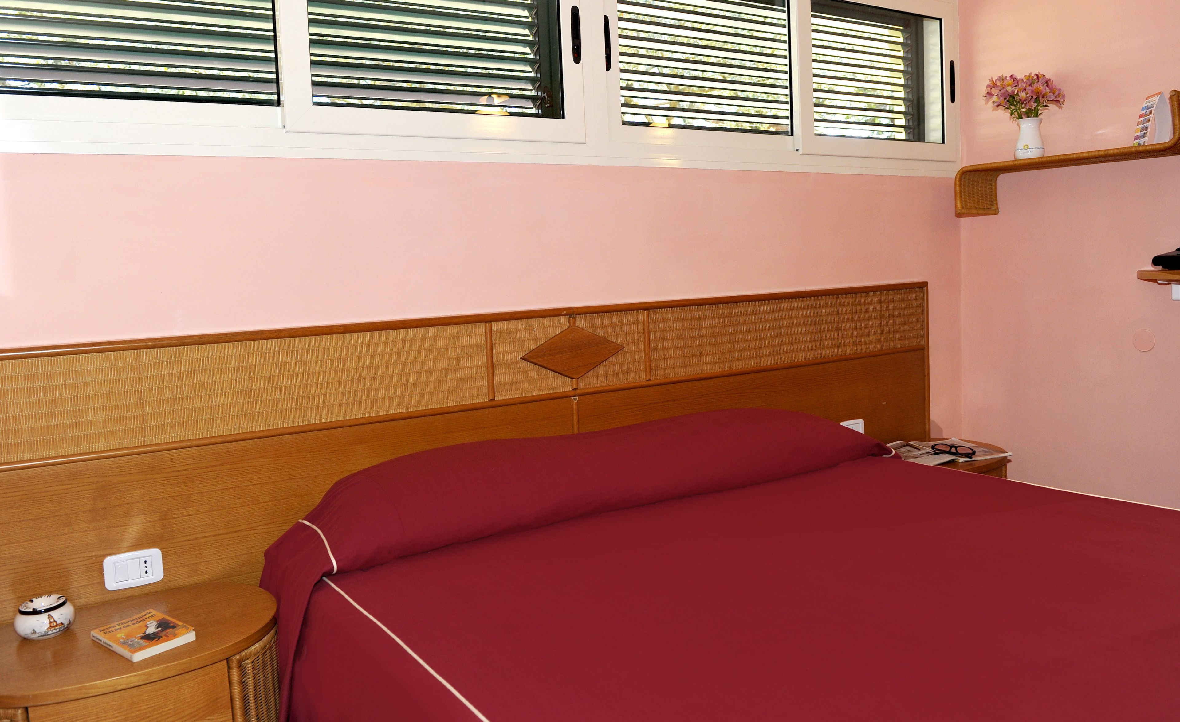 A bedroom with a wooden headboard, a red bedspread, a bedside table with a book and a lamp, and a window with blinds.