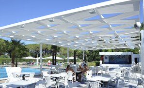 Een zwembadterras met witte parasols, ligbedden en tafels onder een witte pergola.