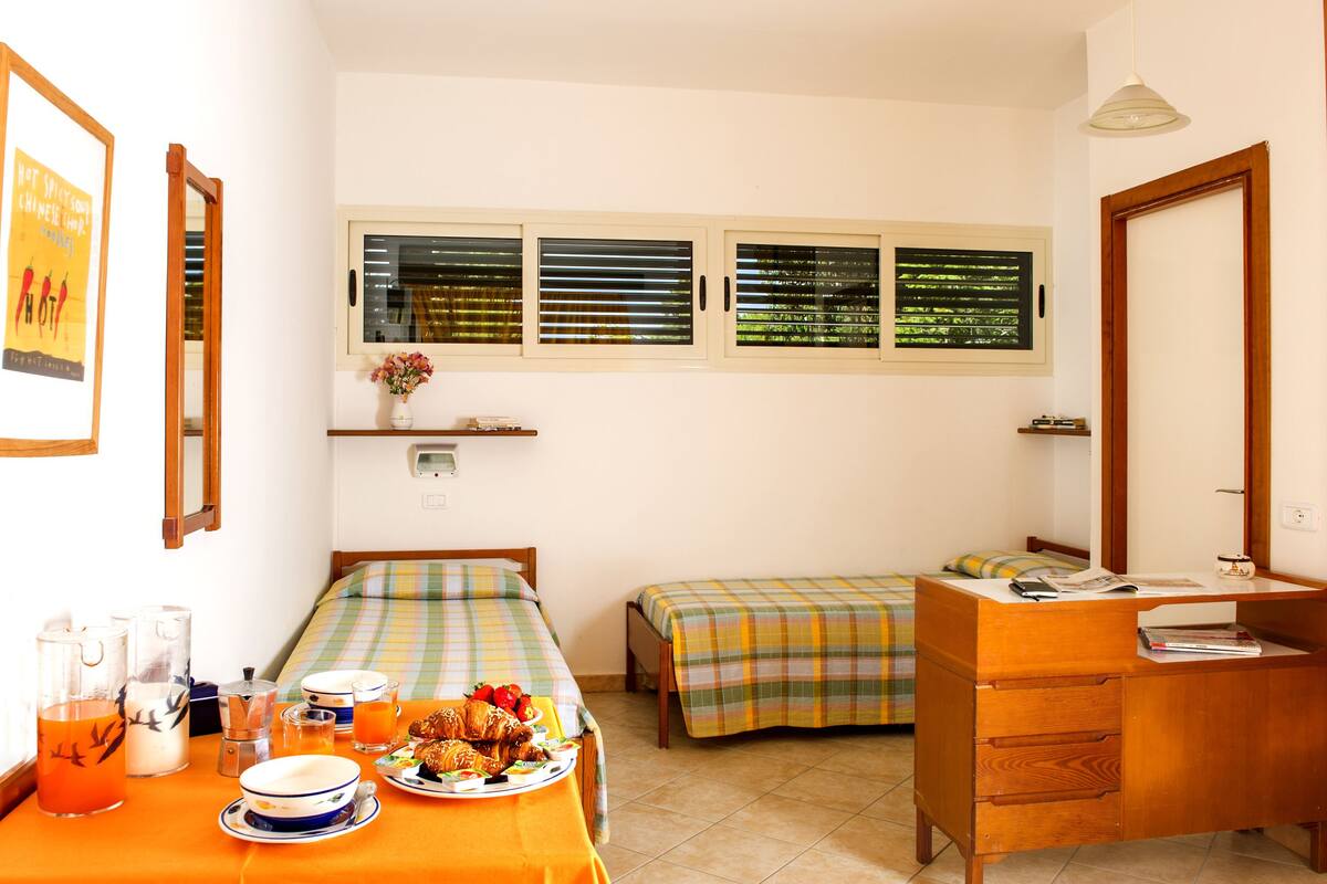 A small, clean room with two beds, a wooden desk, and a window with blinds.