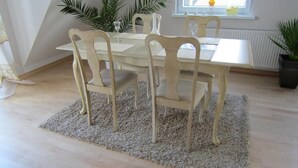 Un coin repas avec une table blanche et quatre chaises, un tapis gris et deux plantes en pot.