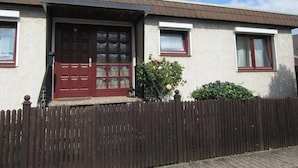 Une maison à deux étages avec une clôture en bois marron, une porte rouge et un petit jardin fleuri.