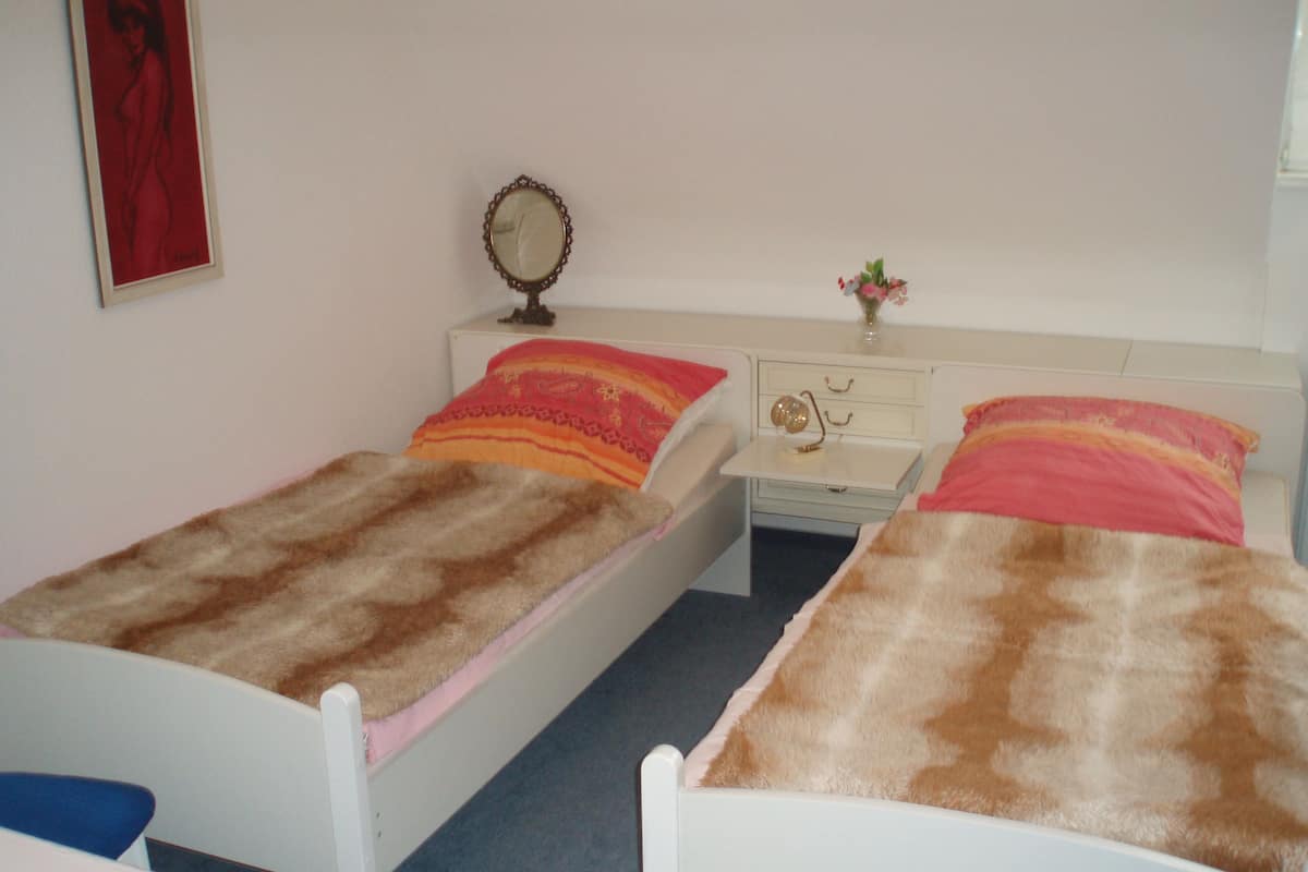 A twin room with two single beds, a bedside table with a mirror, a small table with a lamp, and a vase with flowers.