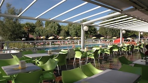 Un área de comedor al aire libre con sillas y mesas verdes, una piscina al fondo y sombrillas que dan sombra.