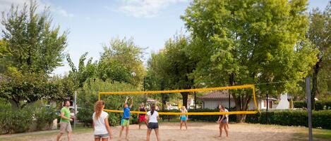 Un grupo de personas jugando voleibol en una cancha de arena rodeada de árboles.
