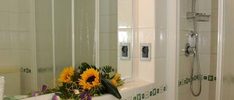 A bathroom with a sink, mirror, shower, and a flower arrangement on the counter.