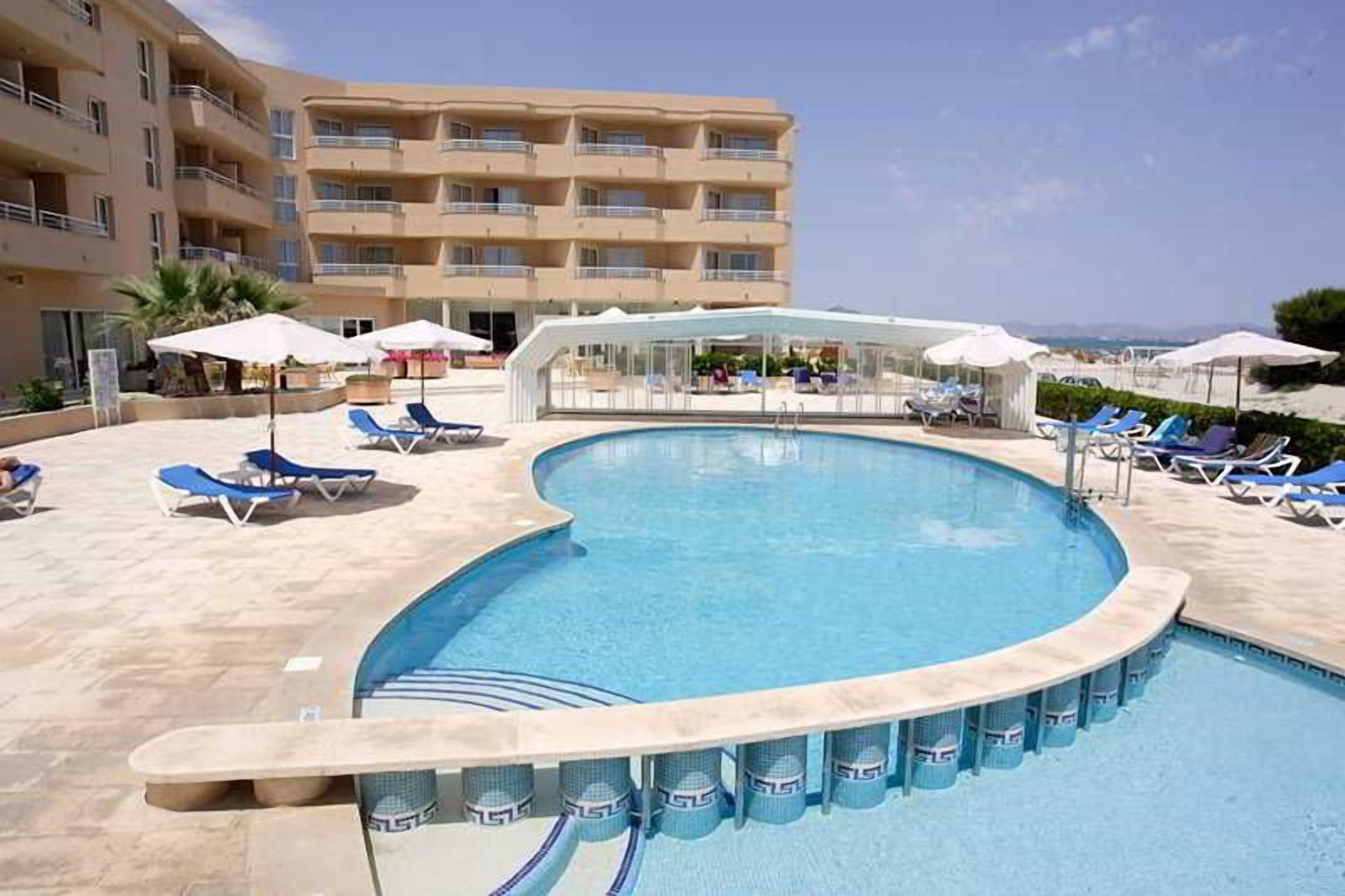 Piscina con sillones de descanso y sombrillas, rodeada por un edificio con balcones.