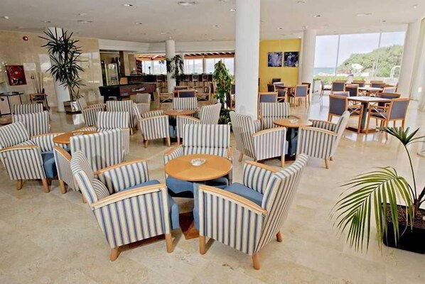 Un vestíbulo de hotel con sillones a rayas, mesas redondas y grandes ventanales que ofrecen una vista.