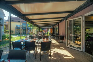 An outdoor dining area with tables and chairs, large windows, and a covered section. - Résidence Les Rives de la Fecht (Ingersheim)