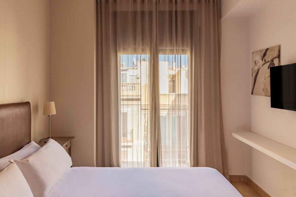 Une chambre d’hôtel avec un lit, une table de chevet avec une lampe, un téléviseur fixé au mur et une fenêtre avec des rideaux.