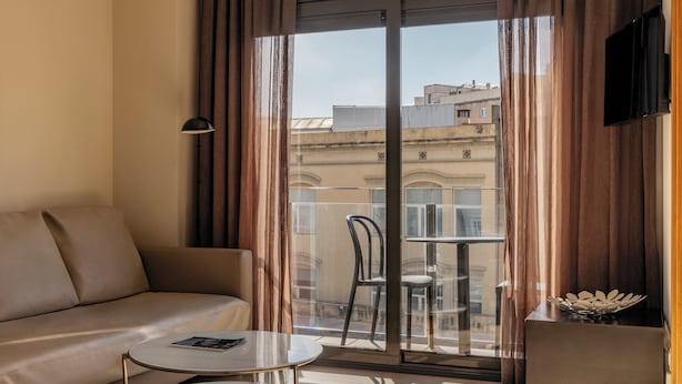 A modern hotel room with a sofa, a small round table, a chair, and a balcony with a view of buildings. - Gran de Gràcia Apartments (Barcelona)