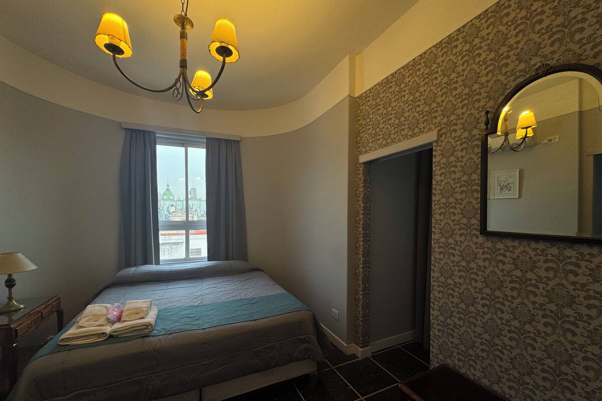 A hotel room with a bed, a window with curtains, a chandelier, a mirror, and patterned wallpaper.