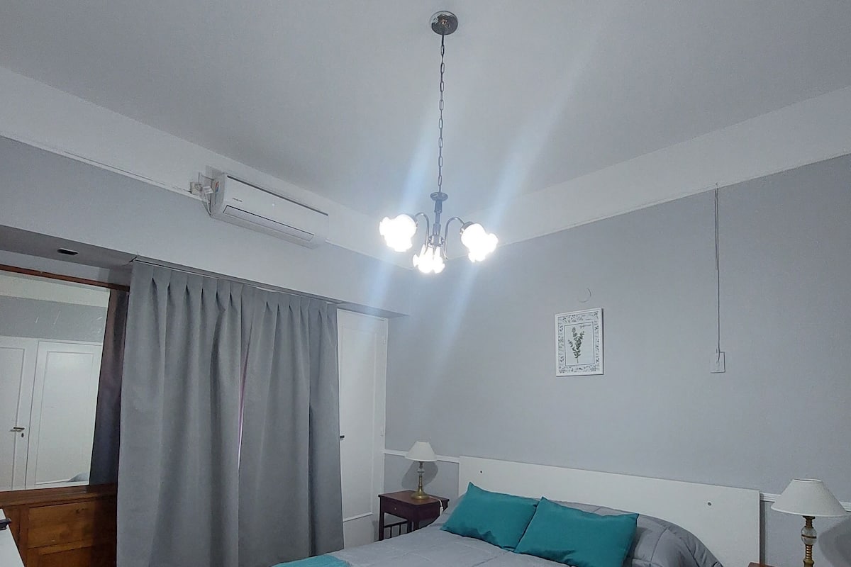 A bedroom with a bed, a nightstand, a lamp, an air conditioning unit, and a window with curtains.