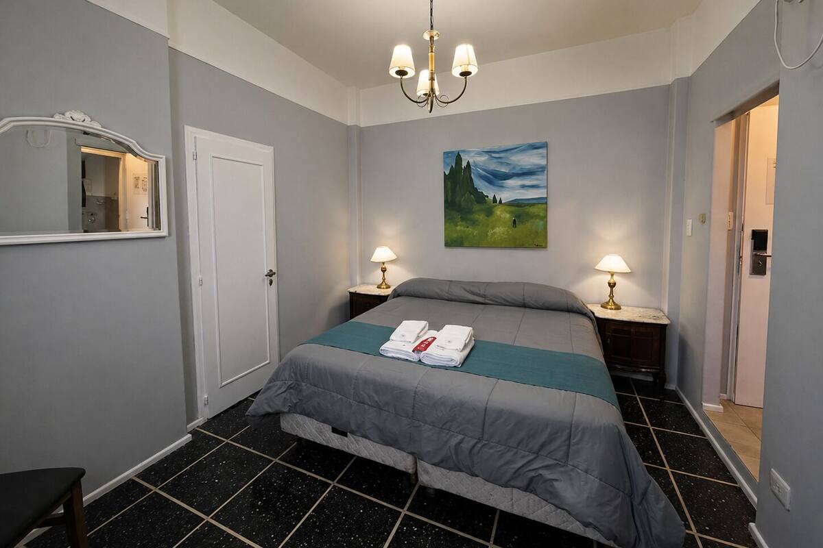A bedroom with a bed, two bedside lamps, a painting on the wall, a mirror, and a door.
