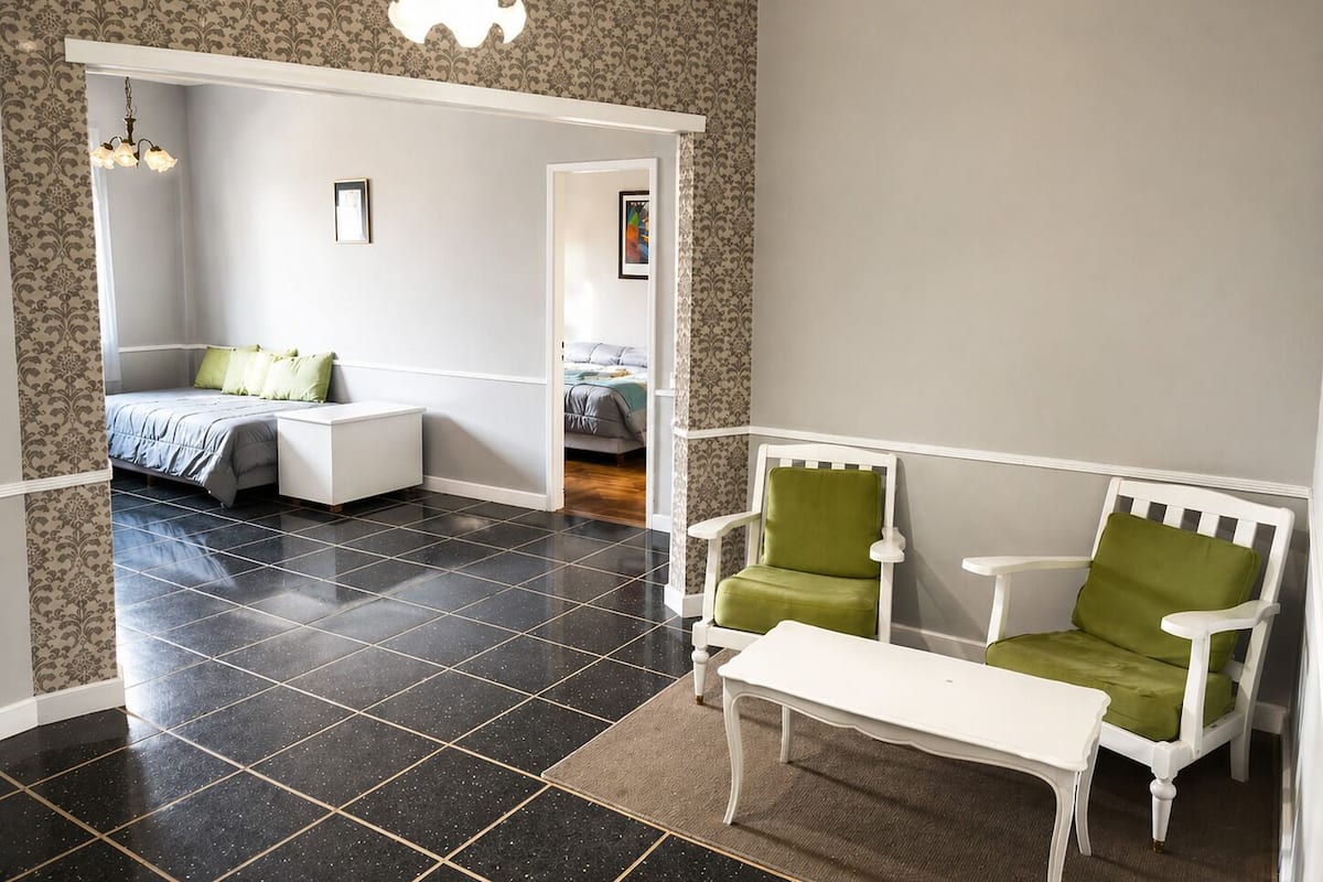 A room with a bed, a white sofa, a green armchair, a white table, and a patterned wallpaper.