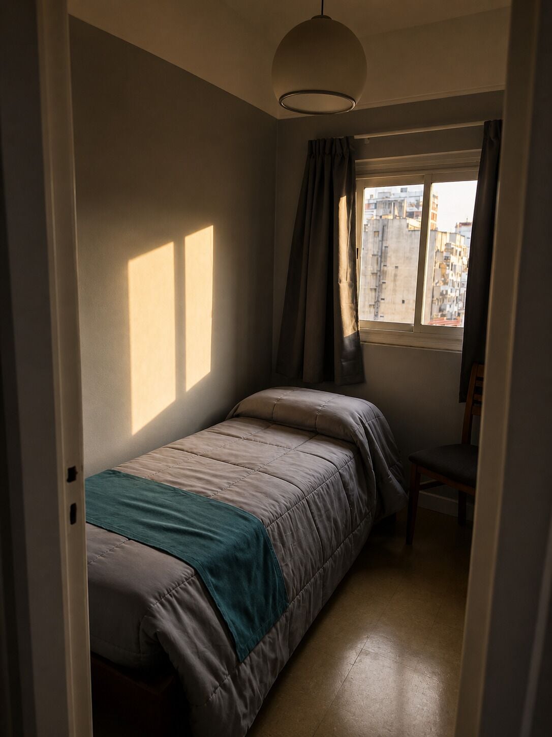 A small, dimly lit room with a single bed, a chair, and a window with curtains.