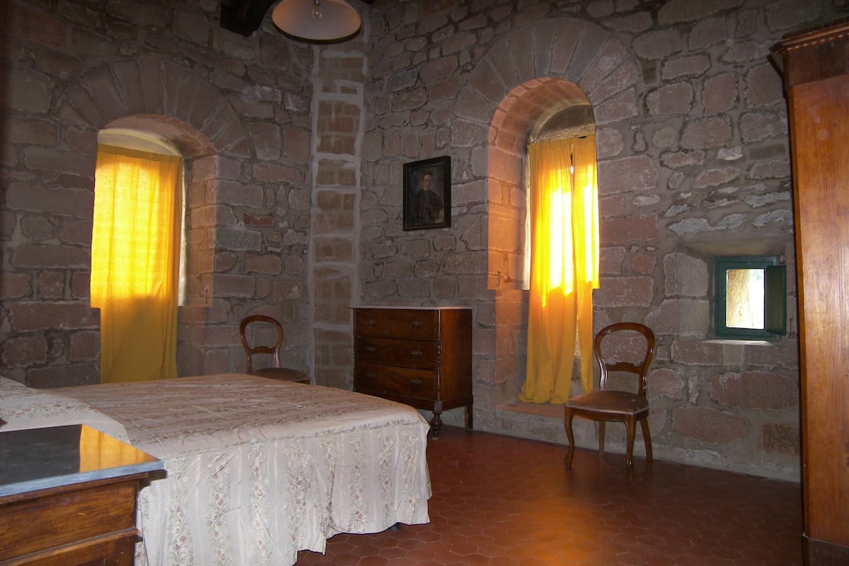 Ein Zimmer mit Steinwänden, einem Bett mit weißer Bettdecke, zwei Holzstühlen, einer Holzkommode und einem kleinen Fenster mit grünem Rahmen.