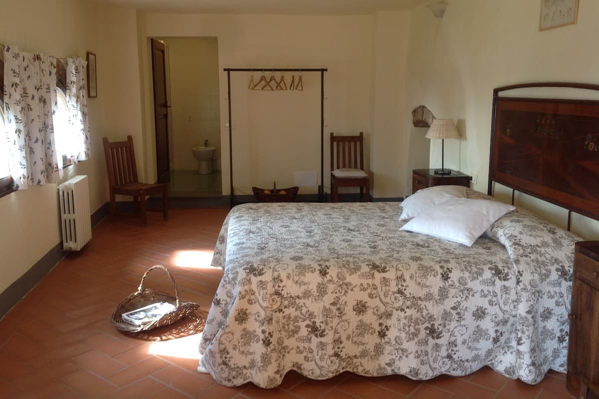Ein Schlafzimmer mit Bett, einem Sessel, einem Heizkörper, einem Fenster mit Vorhängen und einer Tür zu einem Badezimmer.
