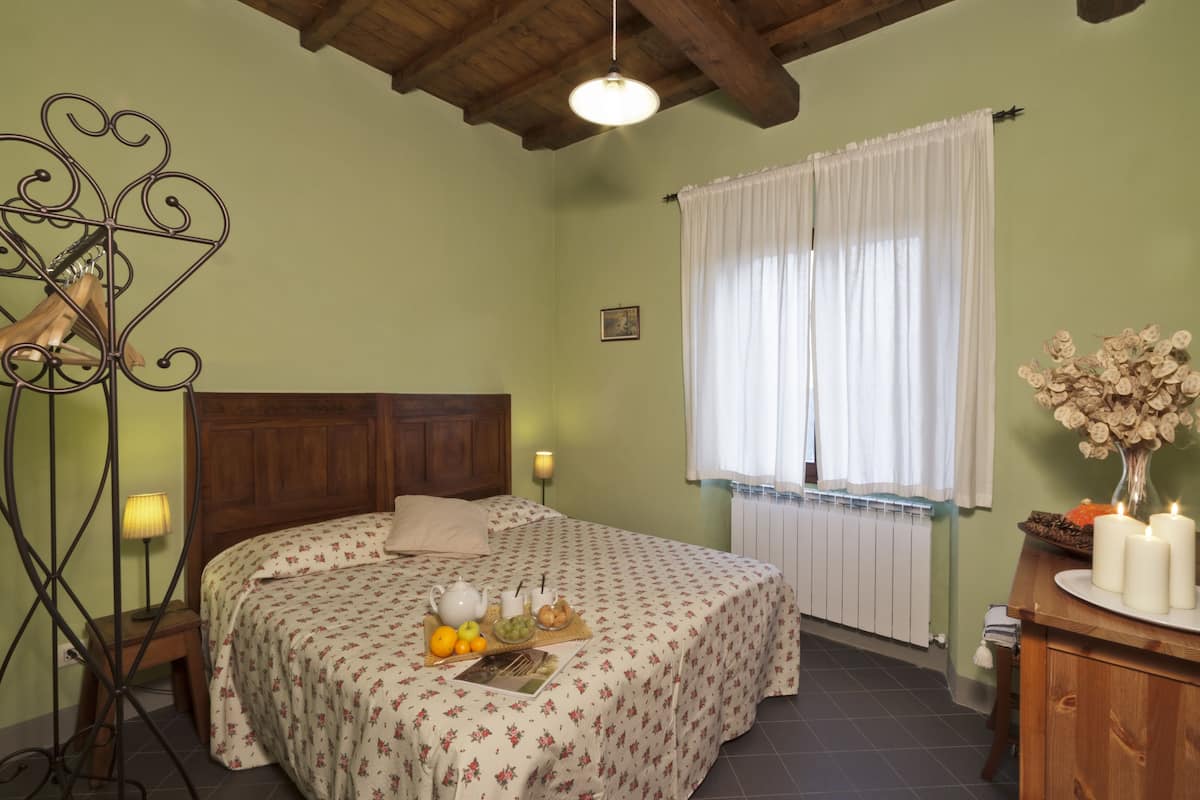 Ein Schlafzimmer mit hölzernem Kopfteil, einem Bett mit floralem Bettzeug, einem Nachttisch mit Lampe, einem Fenster mit weißen Vorhängen und einer Vase mit Trockenblumen auf einer Kommode.