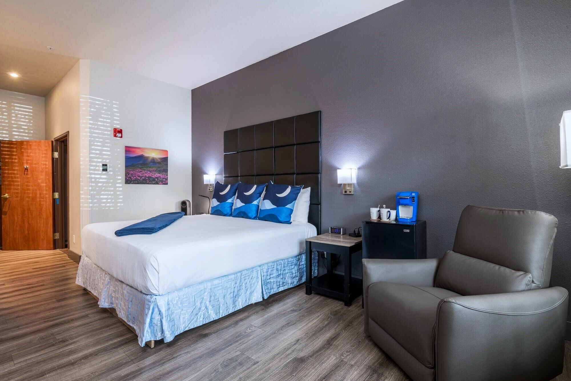 A hotel room with a bed, a chair, a nightstand, and a wall-mounted coffee machine.