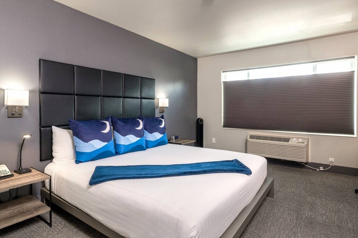 A hotel room with a large bed, a headboard with a built-in panel, two bedside lamps, a window with blinds, and a wall-mounted air conditioning unit.