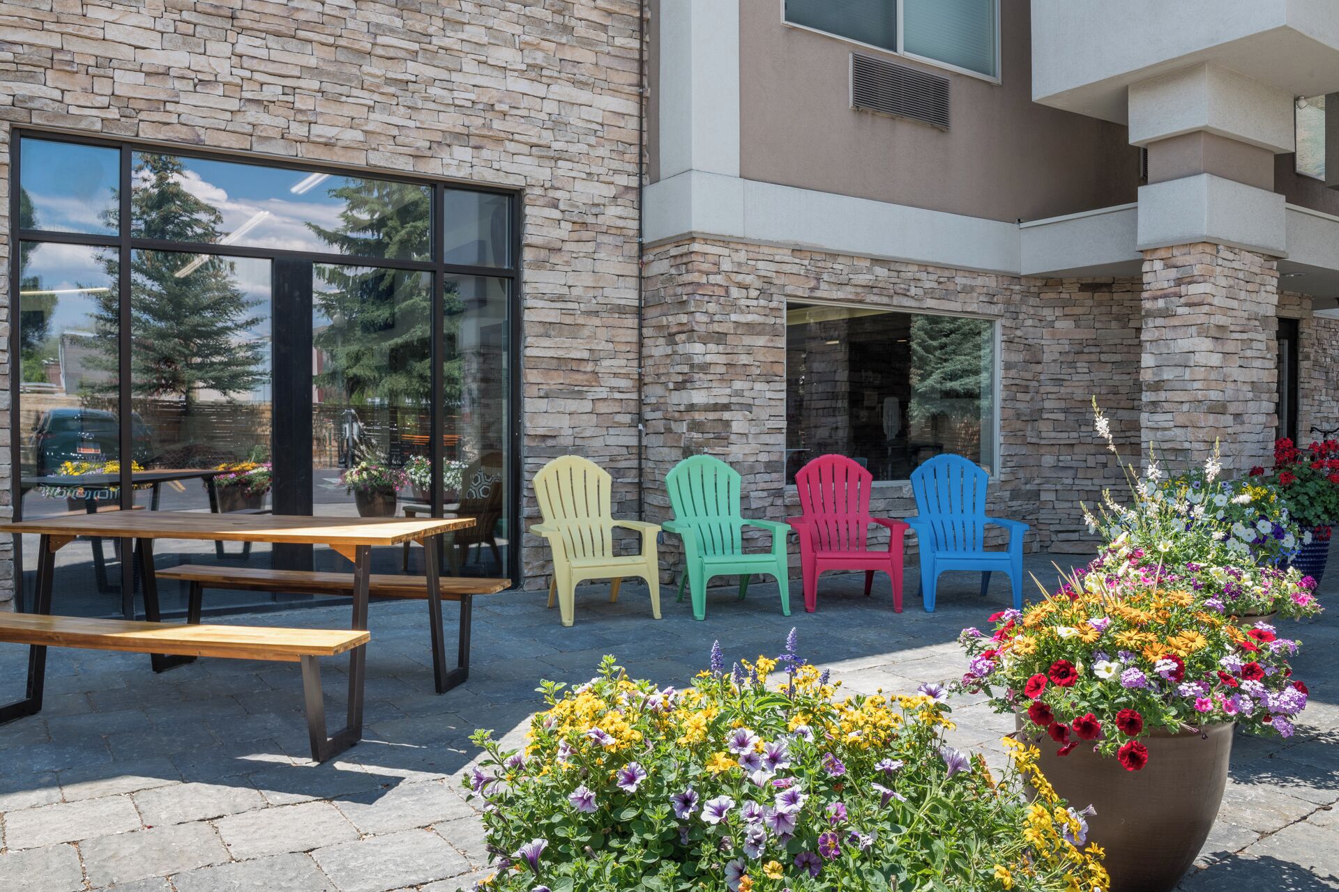 A patio with a stone wall, a wooden table, colorful Adirondack chairs, and potted flowers.