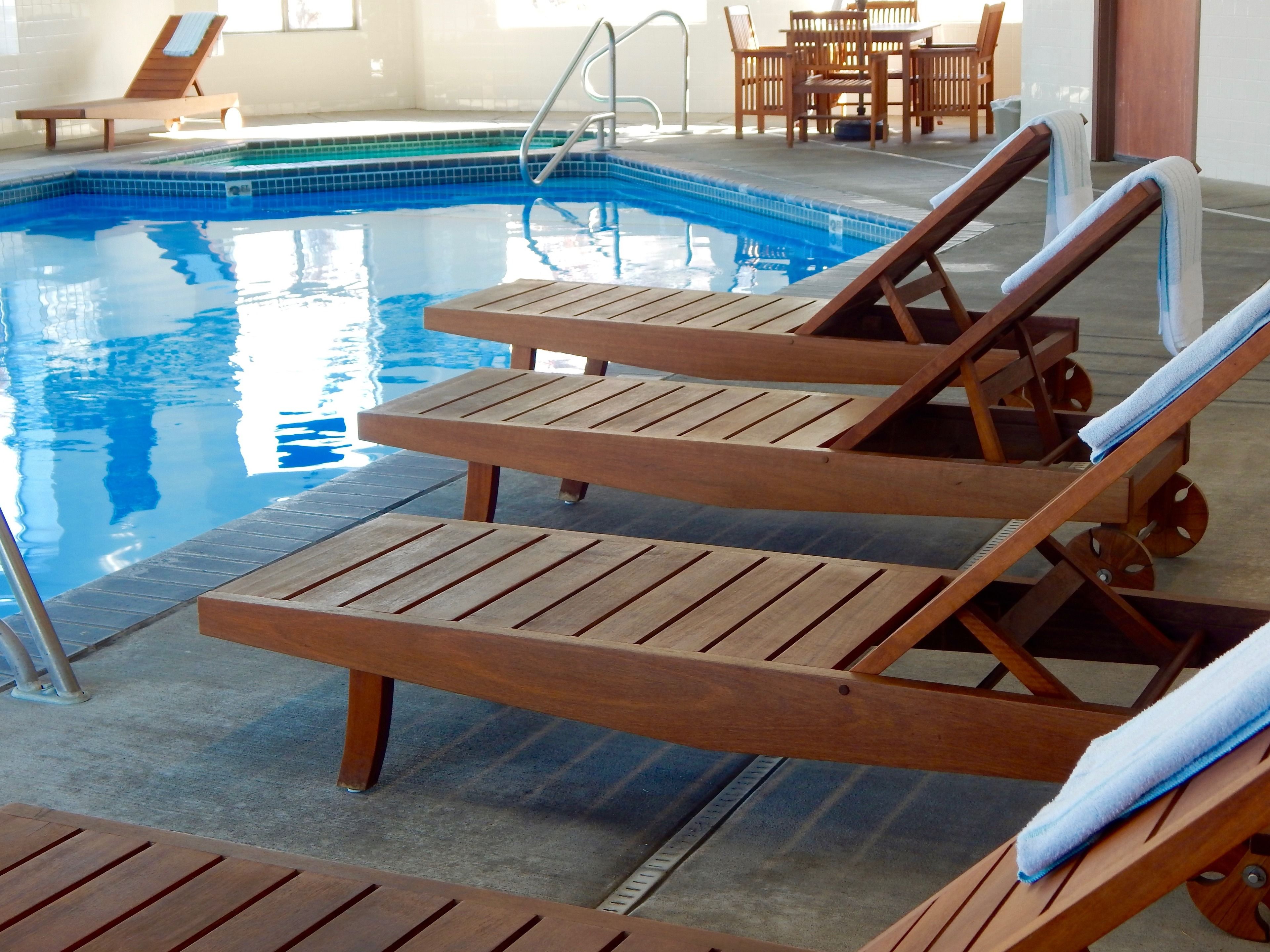 A pool area with wooden lounge chairs and a table, a pool with clear blue water, and a tiled floor.
